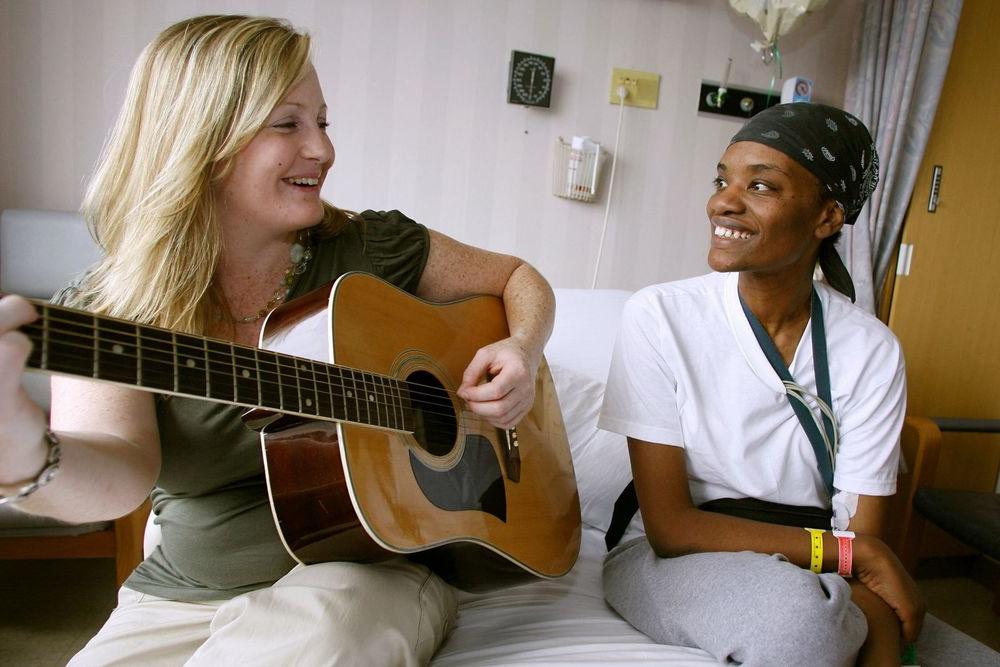 A Music Therapy student sits on a hospital bed next to a patient while playing the guitar.