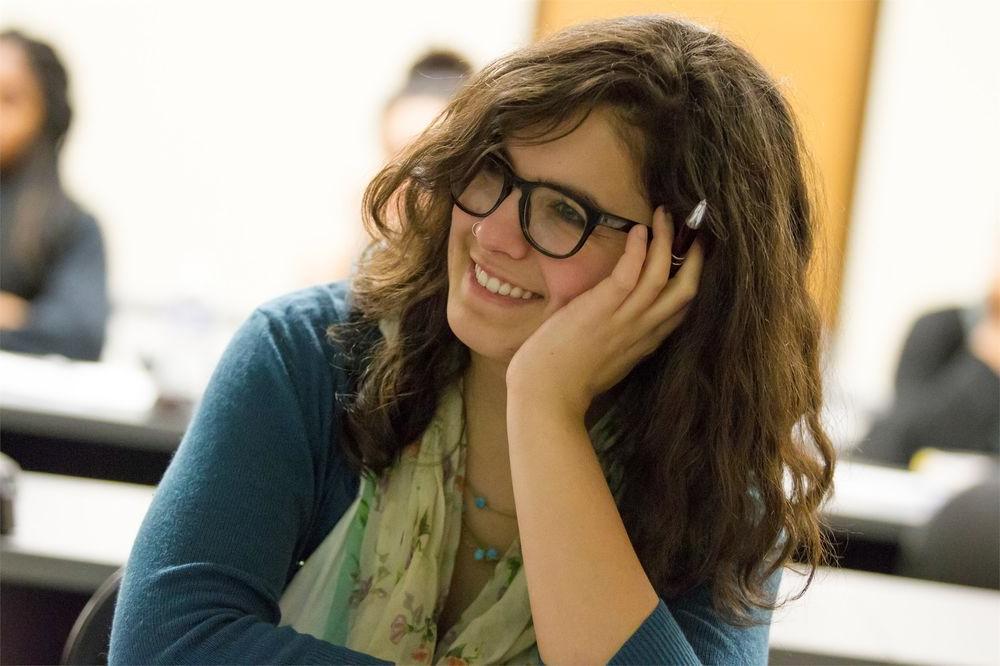 A bespectacled student, her face resting in her left hand, smiles during a class lecture.