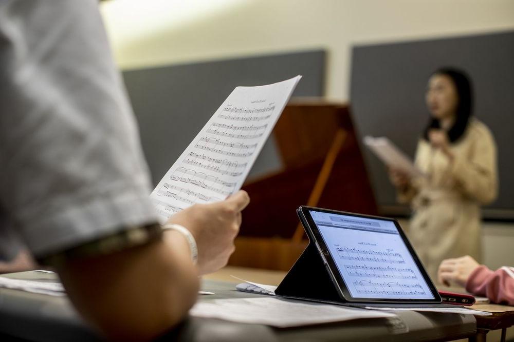 Music students looking at sheet music both on paper and on a computer while a professor lectures at the front of the classroom.