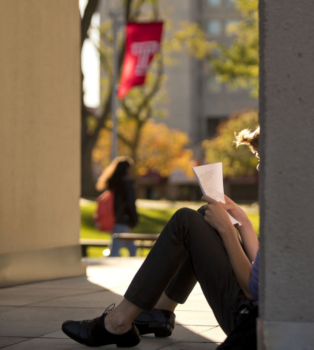 A student sits in the shade on a sunny afternoon, reading a packet of papers